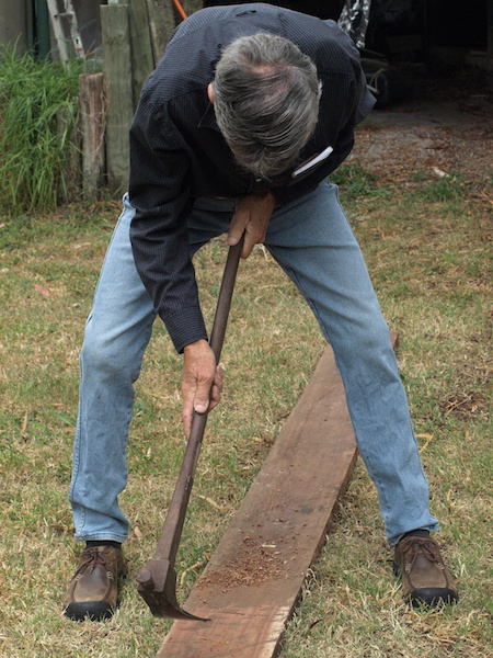 Head down and adzing a rough sawn ironbark plank with a long-handled adze. 