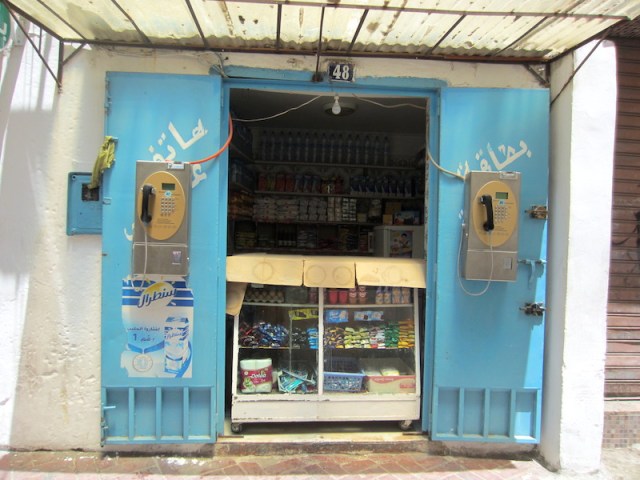 A hole-in-the-wall grocer. Note the two public telephones mounted on the inside of the shop doors. 